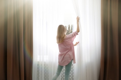 Young woman opening window curtains at home