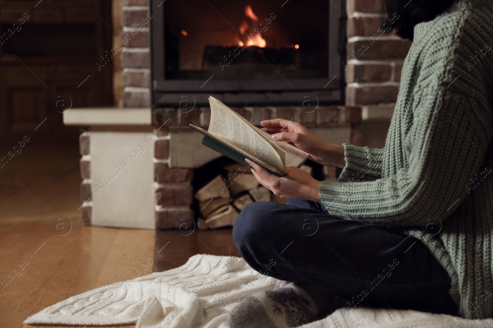 Photo of Woman reading book near burning fireplace at home, closeup