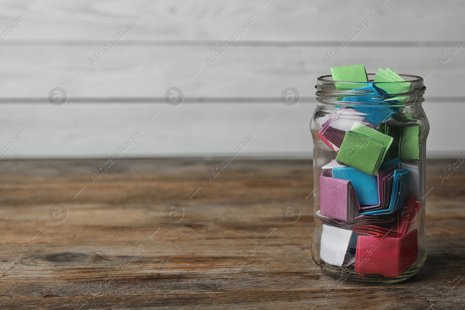 Photo of Glass jar with colorful paper pieces on wooden table, space for text