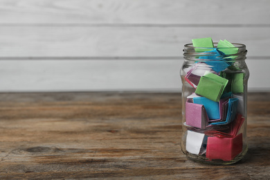Photo of Glass jar with colorful paper pieces on wooden table, space for text