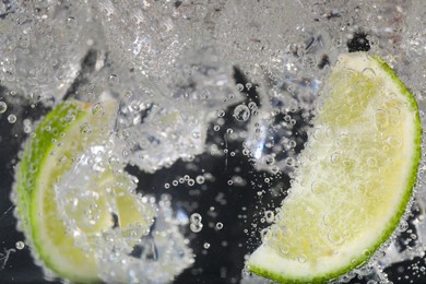 Photo of Juicy lime slices and ice cubes in soda water against black background, closeup