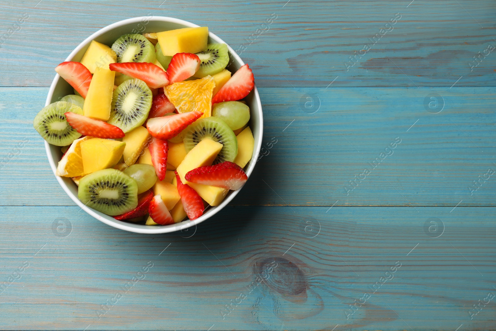Photo of Tasty fruit salad in bowl on light blue wooden table, top view. Space for text