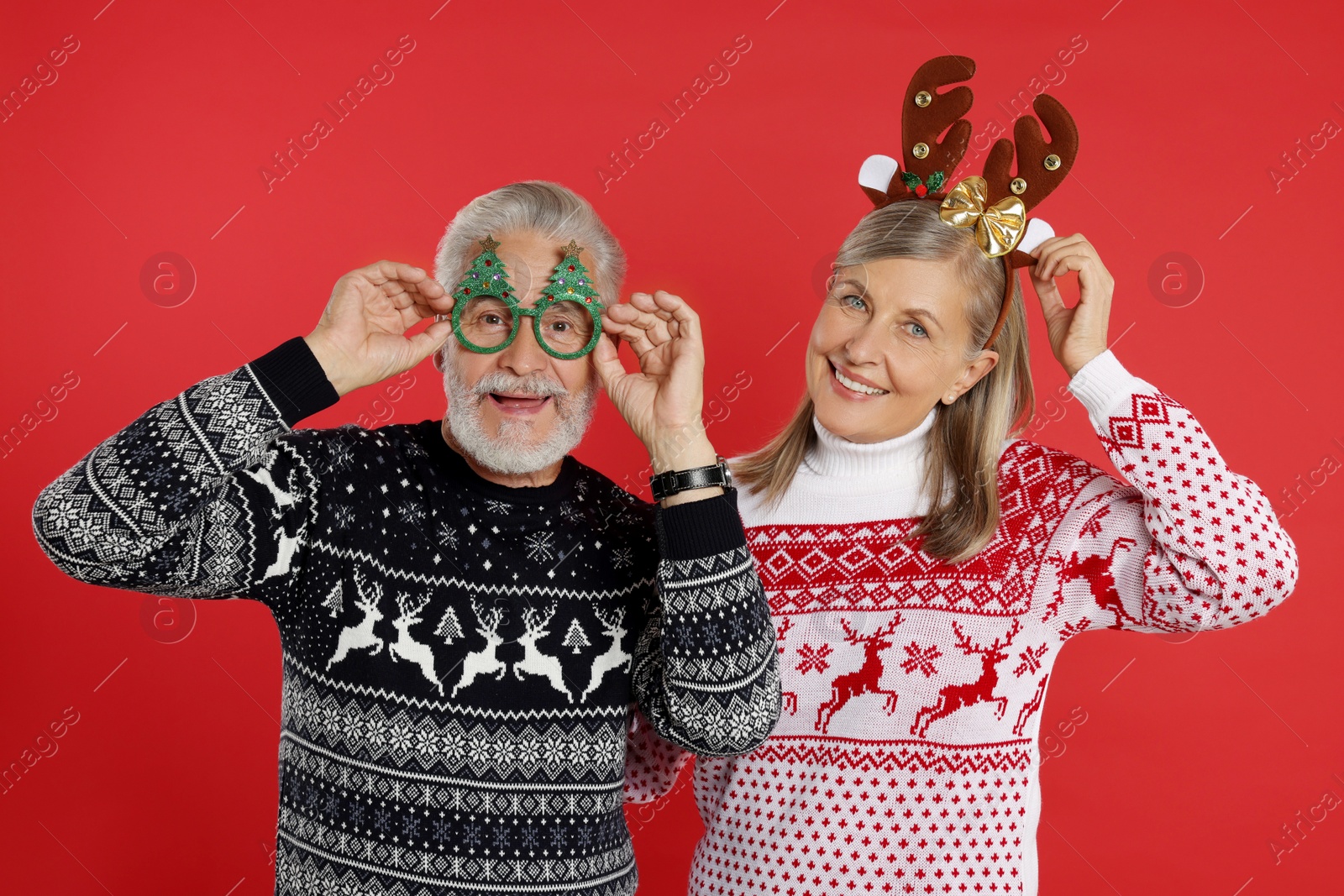 Photo of Happy senior couple in Christmas sweaters, reindeer headband and funny glasses on red background