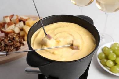 Dipping ham into fondue pot with tasty melted cheese at white table, closeup