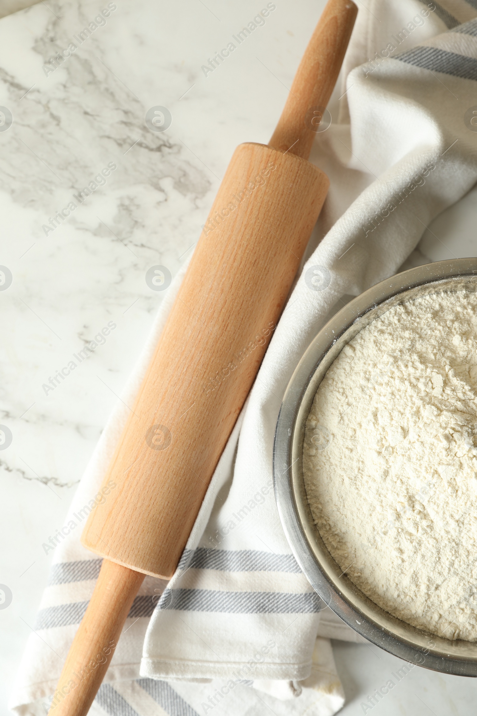 Photo of Flour in bowl, rolling pin and napkin on white marble table, top view