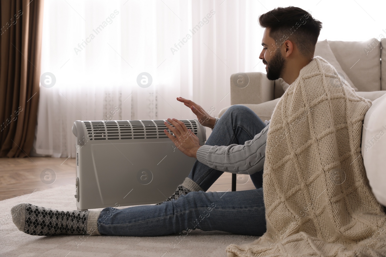 Photo of Young man warming hands near electric heater at home