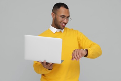 Happy young intern with laptop checking time on light grey background