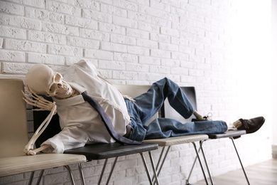 Human skeleton in office wear lying on chairs near brick wall indoors