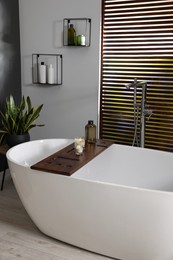 Stylish bathroom interior with ceramic tub, candles and care products on wooden bath tray