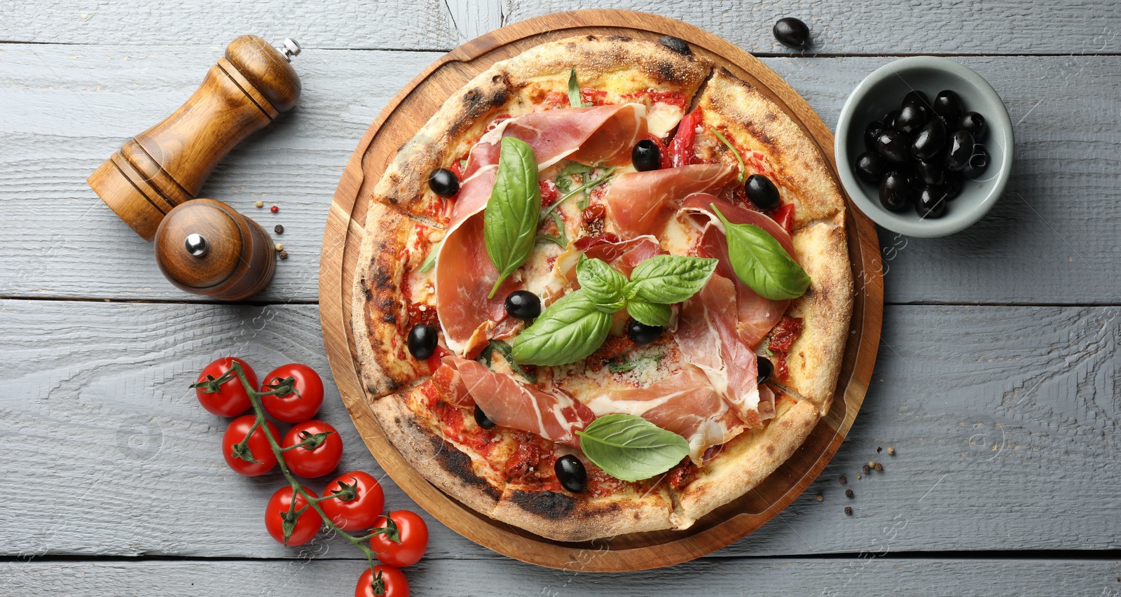 Photo of Tasty pizza with cured ham, olives, tomatoes and basil on gray wooden table, flat lay