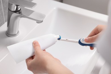 Photo of Woman squeezing toothpaste from tube onto toothbrush above sink in bathroom, closeup