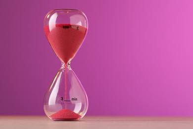 Photo of Hourglass with flowing red sand on table against violet background, space for text