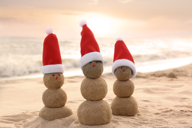 Photo of Snowmen made of sand with Santa hats on beach near sea at sunset. Christmas vacation