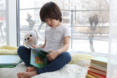 Photo of Cute little boy with toy reading book near window at home