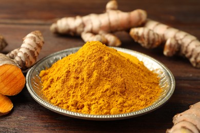 Plate with aromatic turmeric powder and cut roots on wooden table, closeup