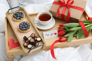 Photo of Tasty breakfast served in bed. Delicious desserts, tea, flowers, gift box and card with phrase I Love You on tray