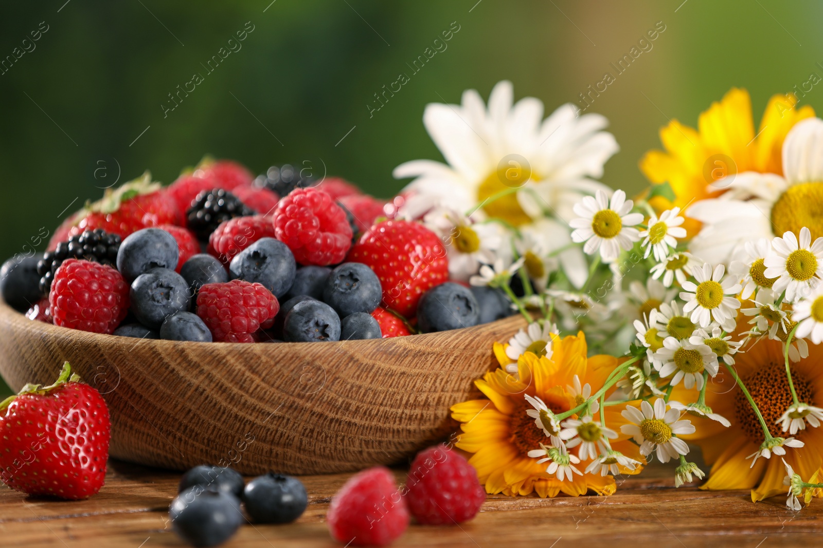 Photo of Bowl with different fresh ripe berries and beautiful flowers on wooden table outdoors