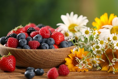 Photo of Bowl with different fresh ripe berries and beautiful flowers on wooden table outdoors