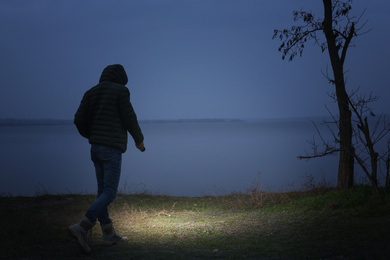 Photo of Man with flashlight walking near river in evening