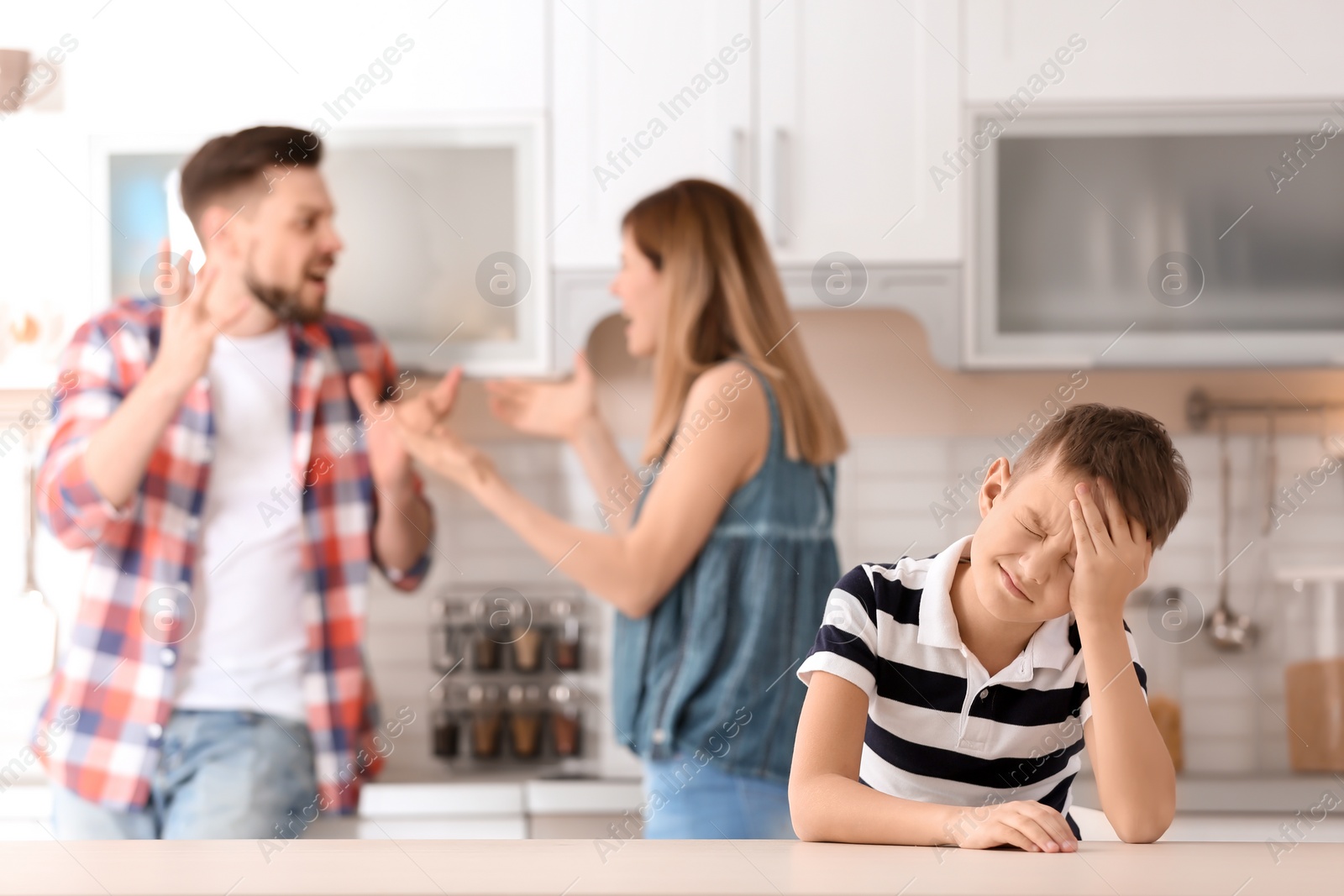 Photo of Little unhappy boy sitting at table while parents arguing on kitchen