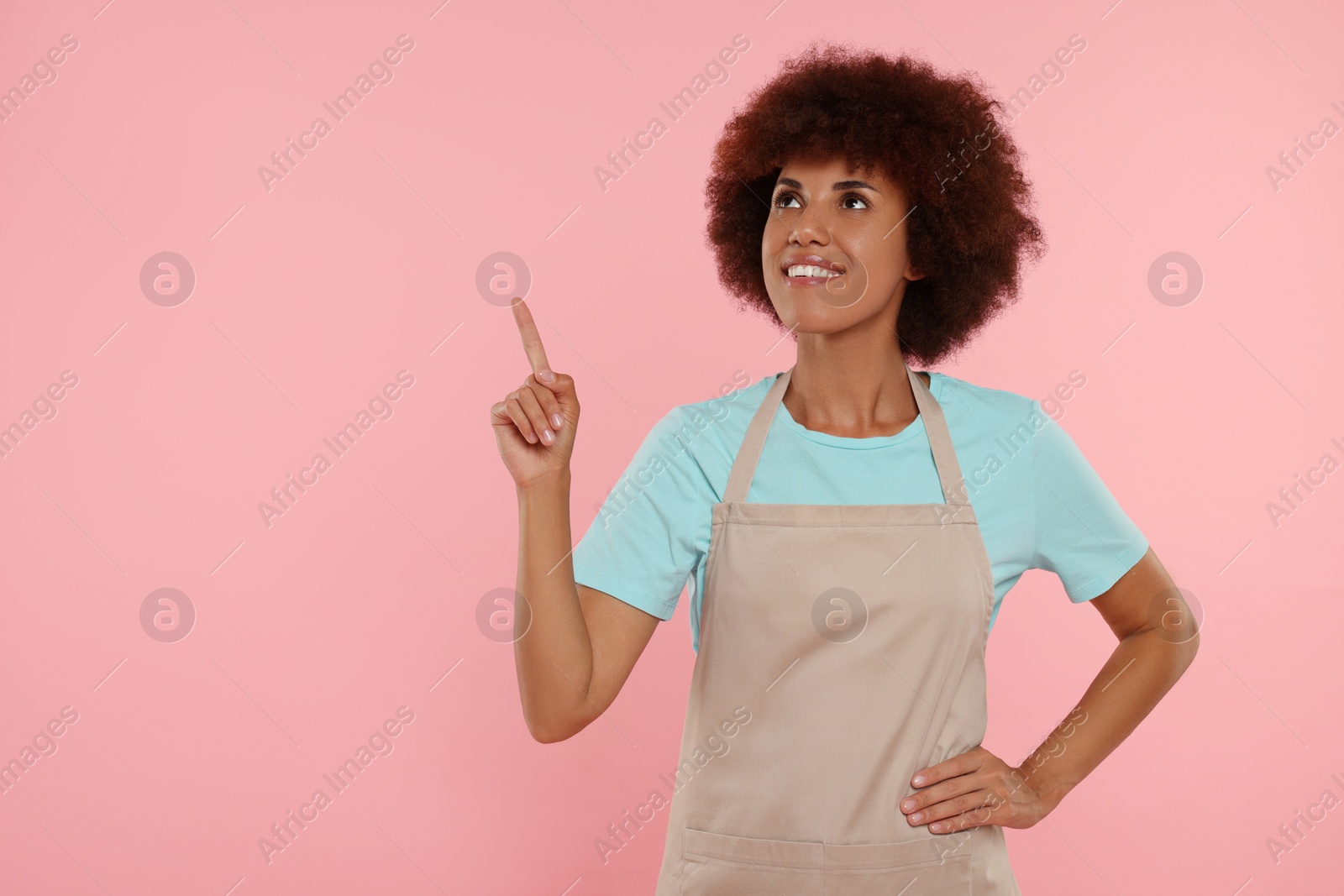 Photo of Happy young woman in apron pointing at something on pink background. Space for text