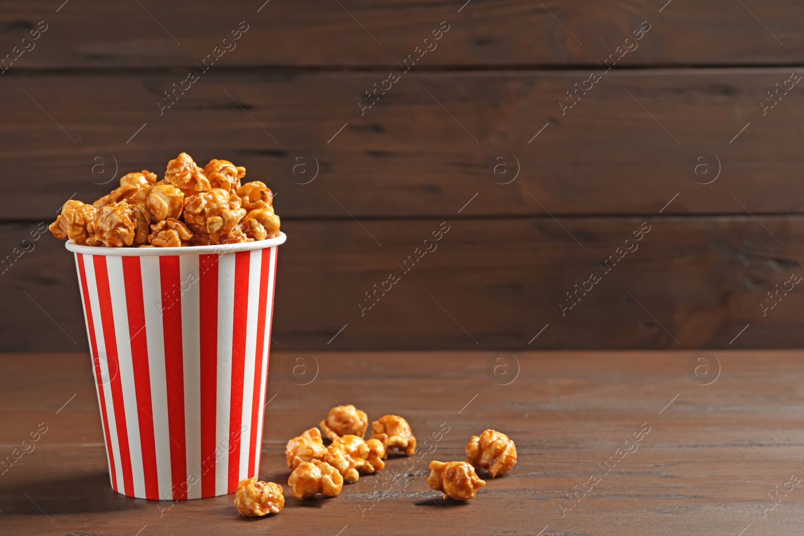 Photo of Delicious popcorn with caramel in paper bucket on wooden table against dark background