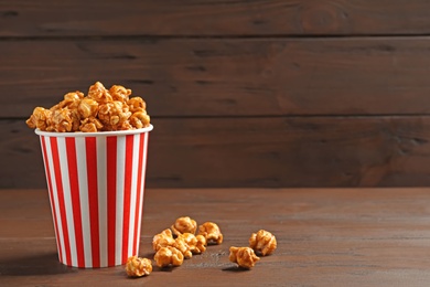 Photo of Delicious popcorn with caramel in paper bucket on wooden table against dark background