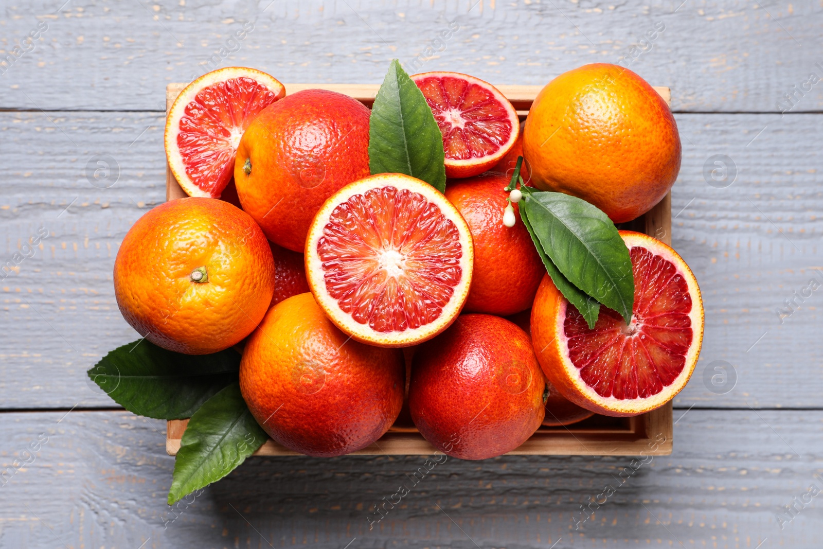 Photo of Crate of ripe red oranges and green leaves on grey wooden table, top view