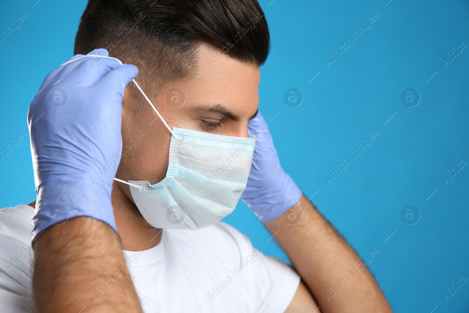 Photo of Man in medical gloves putting on protective face mask against blue background