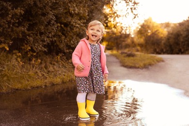 Photo of Little girl wearing rubber boots standing in puddle outdoors. Autumn walk