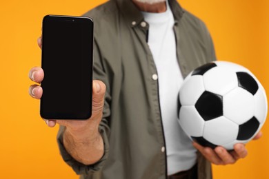 Photo of Sports fan with soccer ball showing smartphone on yellow background, closeup