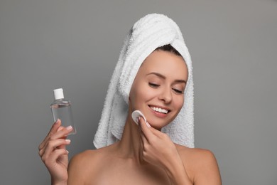 Smiling woman removing makeup with cotton pad and holding bottle on grey background