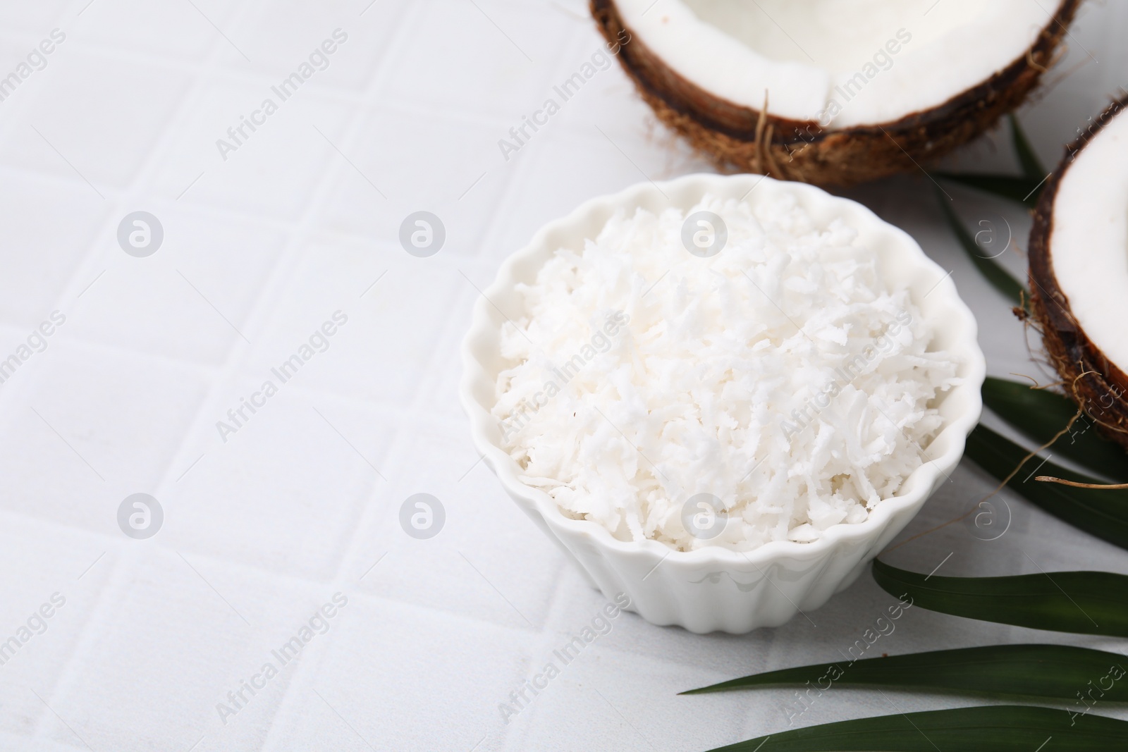 Photo of Coconut flakes in bowl, nuts and palm leaf on white tiled table, space for text