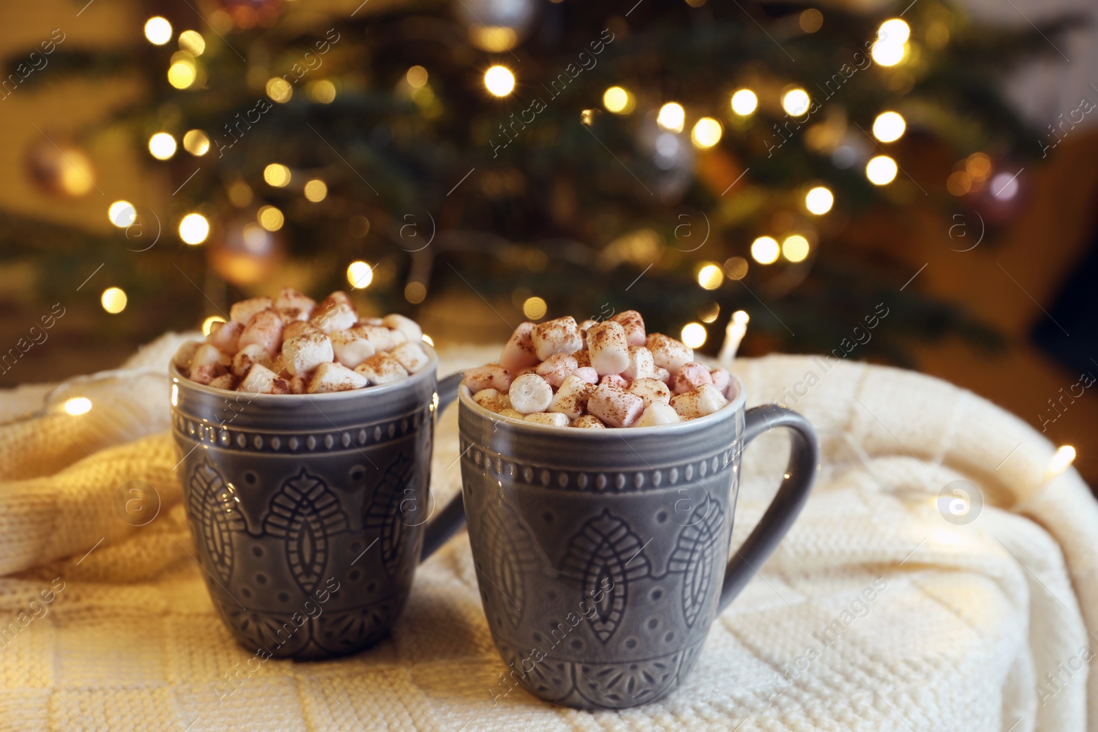 Photo of Cups of tasty cocoa with marshmallows on knitted plaid near Christmas tree indoors