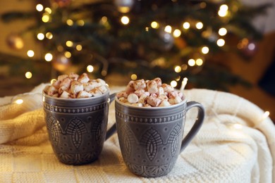 Cups of tasty cocoa with marshmallows on knitted plaid near Christmas tree indoors
