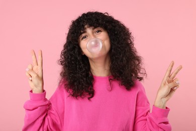 Beautiful young woman blowing bubble gum and showing peace gesture on pink background