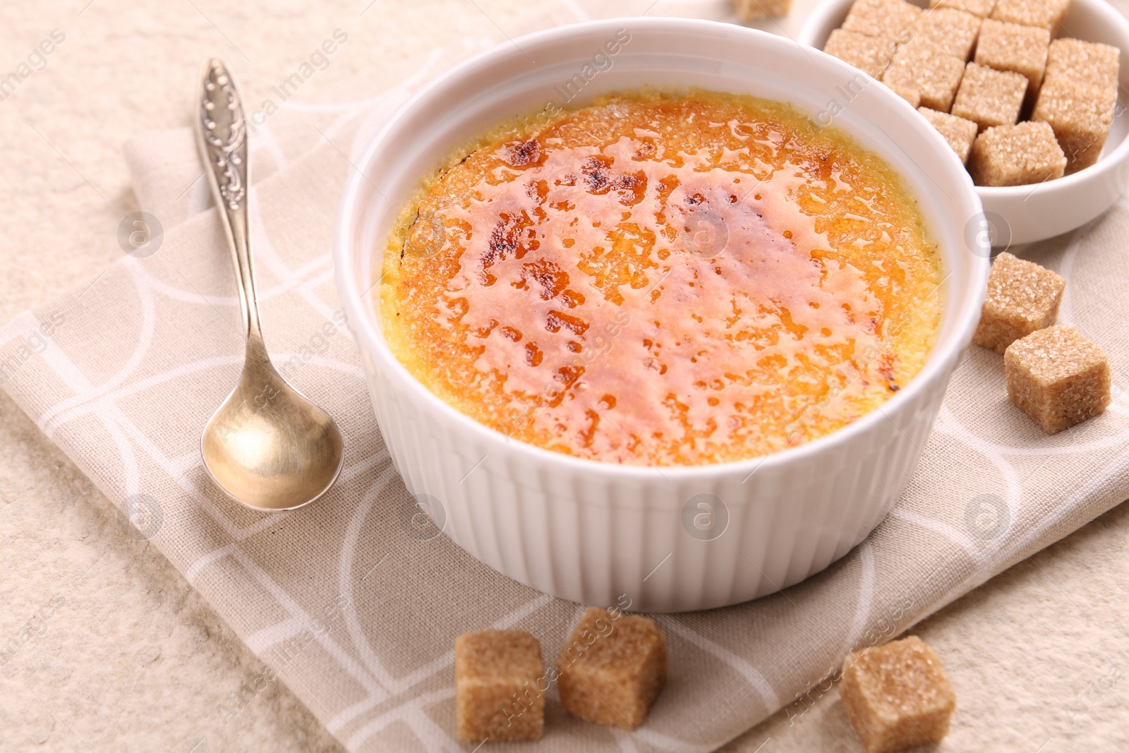 Photo of Delicious creme brulee in bowl, sugar cubes and spoon on light textured table, closeup