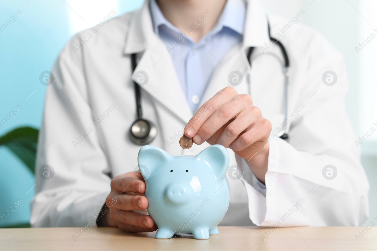 Photo of Doctor putting coin into piggy bank at wooden table, closeup