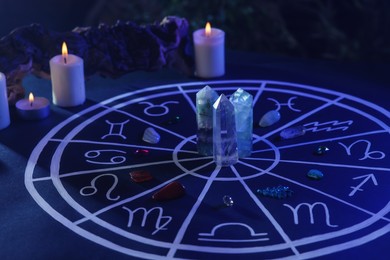Photo of Natural stones for zodiac signs, drawn astrology chart and burning candles on dark blue table