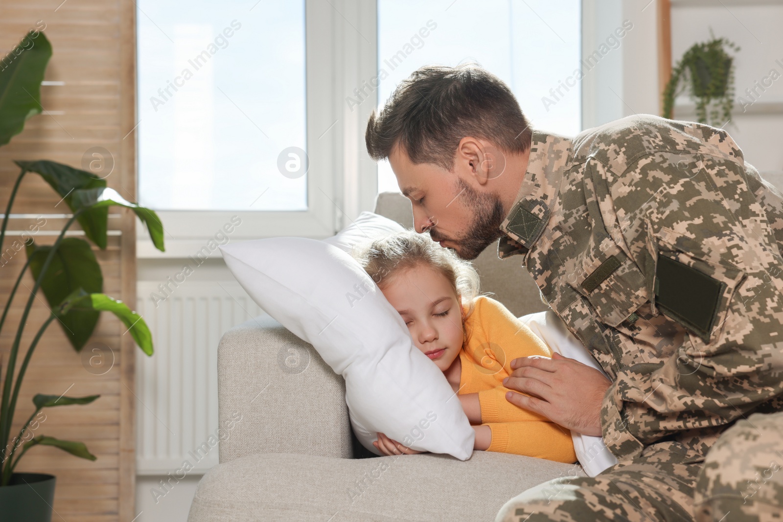 Photo of Soldier in Ukrainian military uniform kissing his daughter while she sleeping on sofa at home. Family reunion