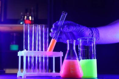Scientist working with laboratory glassware of luminous liquids at table against dark background, closeup