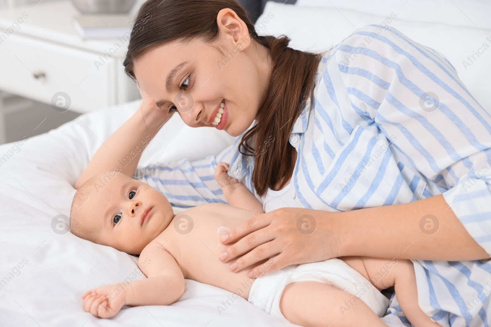 Photo of Happy young woman applying body cream onto baby`s skin on bed