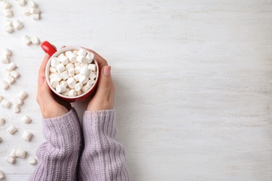 Woman holding cup of tasty cocoa with marshmallows on white wooden table, top view. Space for text