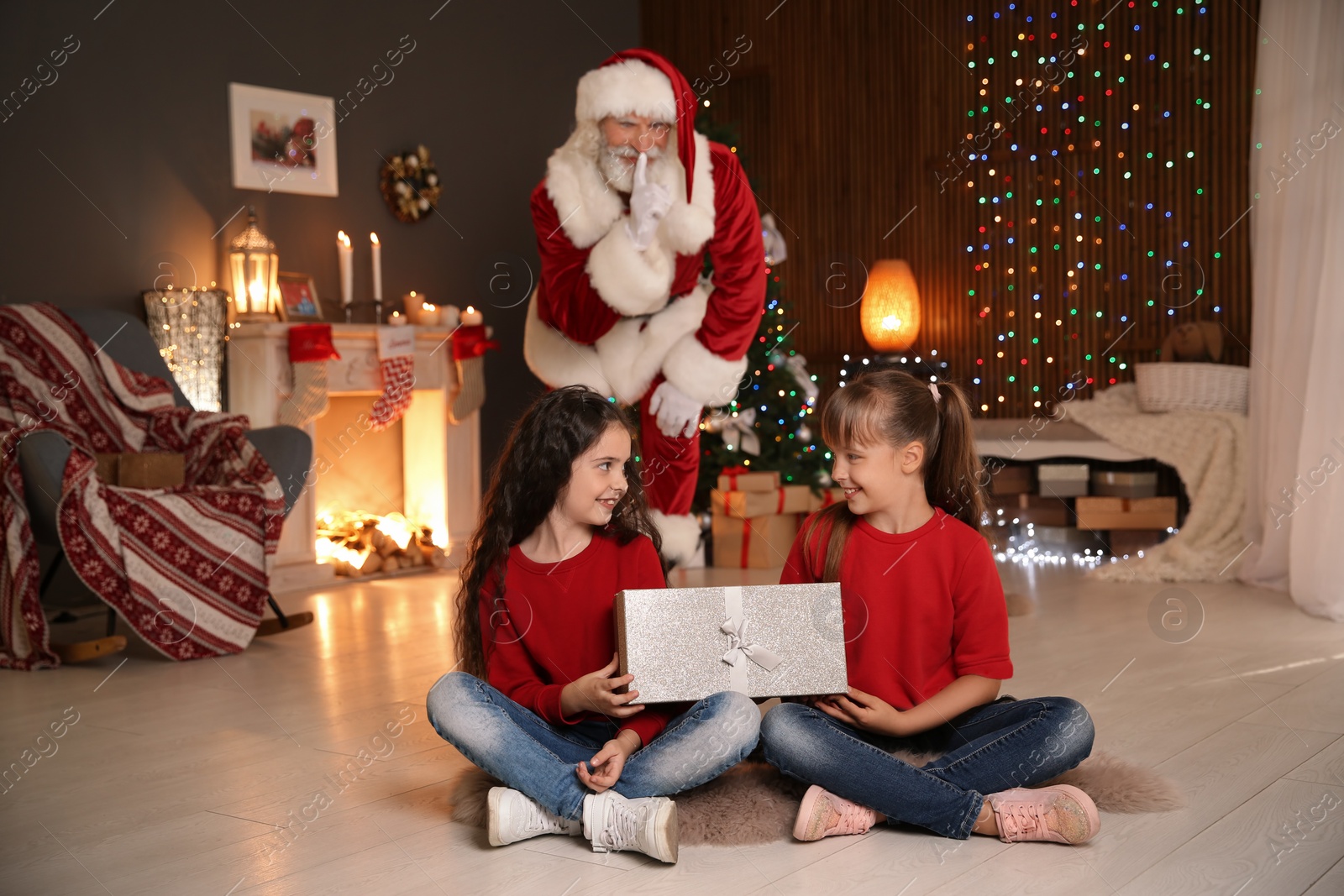 Photo of Little children with Christmas gift and Santa Claus hiding behind them at home