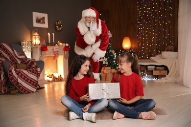 Little children with Christmas gift and Santa Claus hiding behind them at home