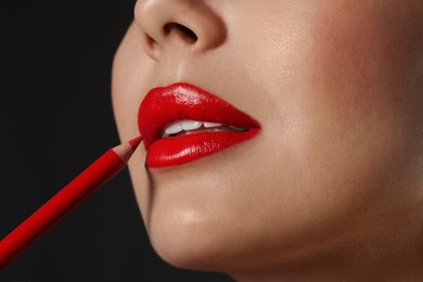 Photo of Young woman applying beautiful red lip pencil on black background, closeup