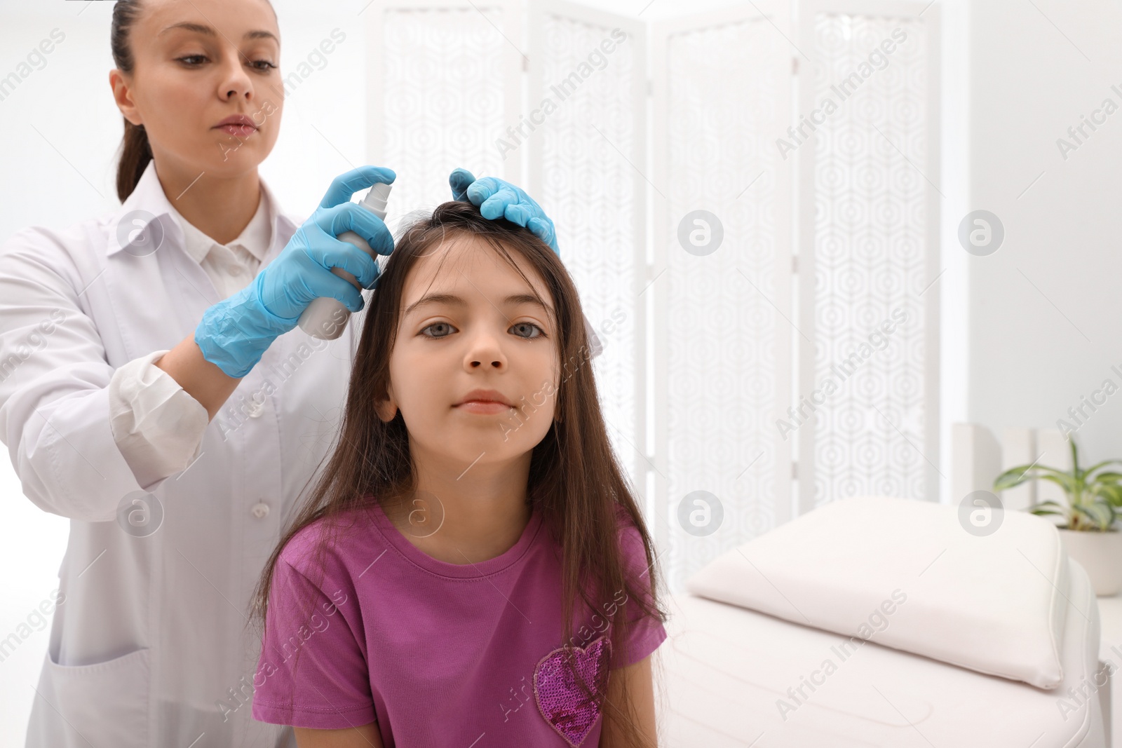 Photo of Doctor using lice treatment spray on little girl's hair indoors