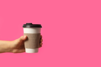 Woman holding takeaway paper coffee cup with cardboard sleeve on pink background, closeup. Space for text