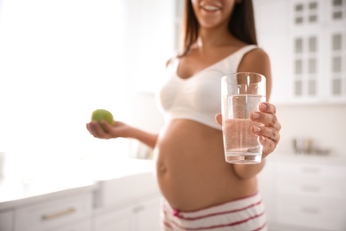Photo of Young pregnant woman with glass of water and apple in kitchen, focus on drink. Taking care of baby health