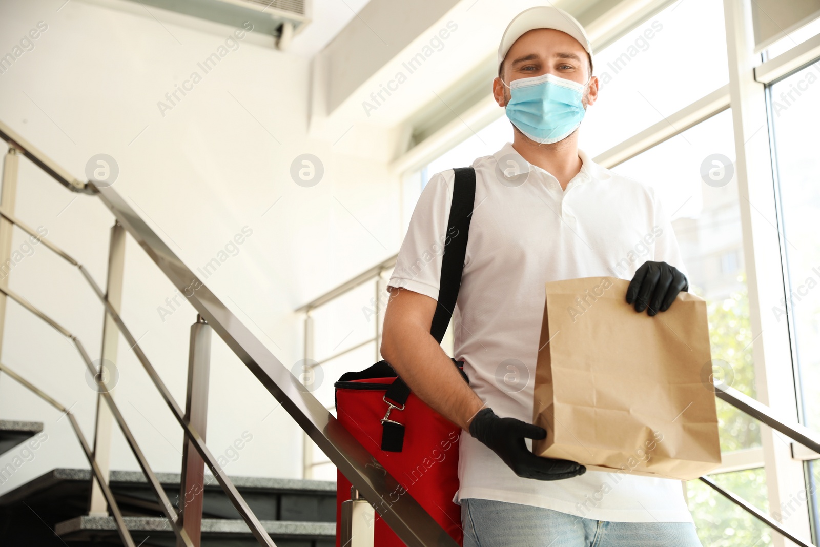 Photo of Courier in protective mask and gloves with order indoors. Restaurant delivery service during coronavirus quarantine